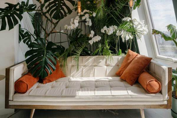 Vertical shot of light cozy boho style living room with modern beige sofa and orange pillows in relaxing area, decorated with different green plants